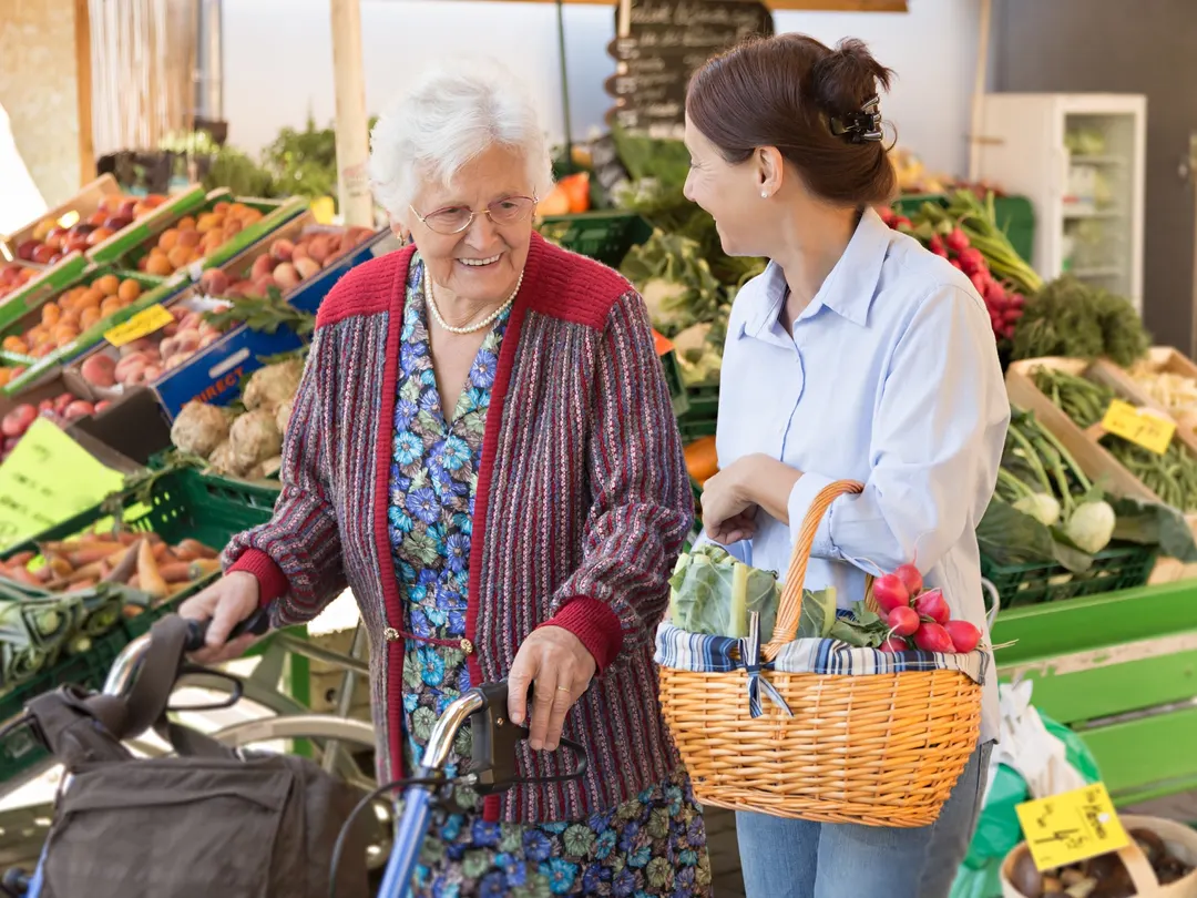 Eine ältere Dame mit Rollator und ihre Begleiterin mit Einkaufskorb auf dem Wochenmarkt.
