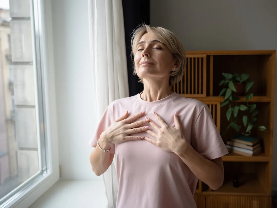 Eine Frau steht mit geschlossenen Augen und den Händen auf der Brust am Fenster und atmet durch.